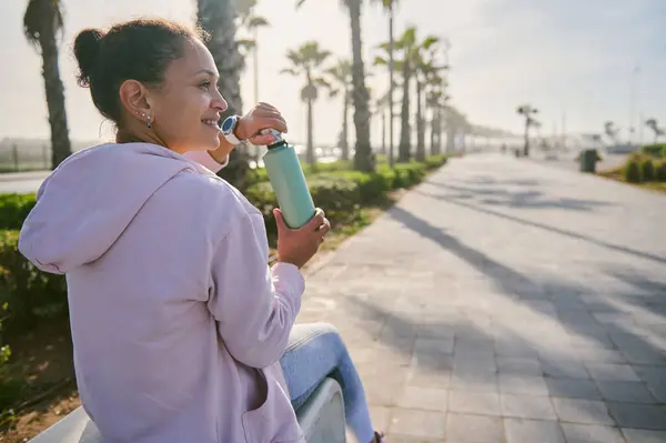从后面看去 一个笑着的女运动员早上在人行道上慢跑后喝水 大家好积极健康的生活方式概念 图库图片