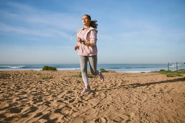 早上在沙滩上慢跑时 身材匀称 体形匀称 体态健壮的年轻女子正在燃烧热量 有吸引力的有决心的女运动员 穿着运动服的跑步者 喜欢户外运动 图库照片