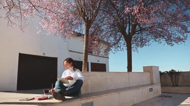 一个美丽的女人坐在盛开的樱桃树下 画着水彩画 在一个美丽的阳光明媚 多风的春日里 有创意的人Hobby 视觉艺术 — 图库视频影像