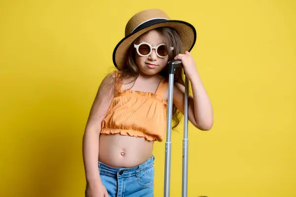 照片上的漂亮小女孩戴着太阳镜 穿着夏装 提着她的手提箱摆姿势 独立在黄色的工作室背景下 旅行和旅游概念 临时空间 图库图片