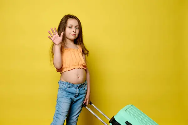 可爱的小女孩背着蓝色的手提箱 用手挥手打招呼 看着相机 隔离在黄色的工作室背景中 复制广告空间 免版税图库照片