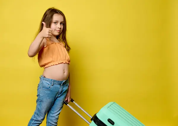 白人可爱的小孩儿 穿着夏装的旅游女孩 看着相机的大拇指姑娘 提着塑料时髦的手提箱 周末出游 与世隔绝的黄色工作室背景 图库图片
