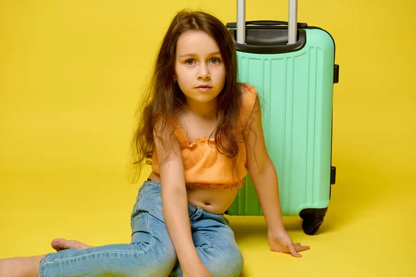 真实的工作室肖像白种人漂亮的小女孩 坐在她时髦的手提箱旁边 穿着夏装 自信地看着相机 孤立的黄色背景 免版税图库照片
