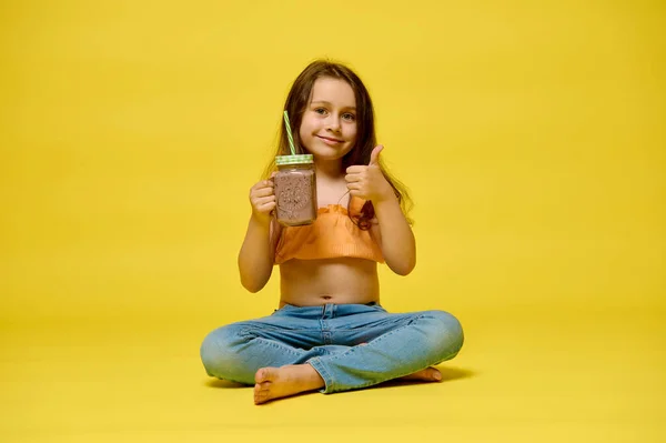 情感肖像白种人小女孩大拇指向上 显示出美味的新鲜挤压果汁 孤立在黄色工作室的背景 大家好休闲活动 暑假的概念 免版税图库图片