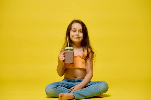 穿着夏装笑着的小女孩 坐在荷花上摆姿势 用稻草喝着玻璃瓶里的鸡尾酒 隔离在黄色工作室的背景下 复制广告空间 图库图片