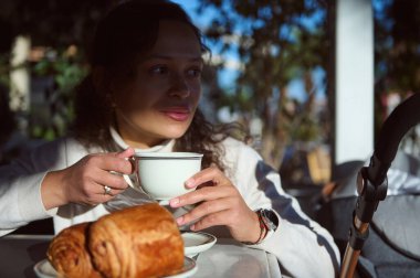 Güzel, düşünceli genç bir kadının kendine güveni tam portresi. Kahvesini açık sokak kafesinde yapıyor, taze demlenmiş espresso içiyor ve lezzetli kızarmış Fransız kruvasanı yiyor.