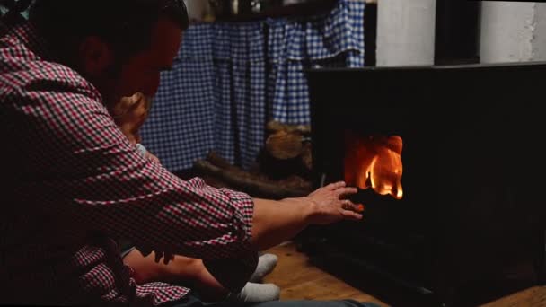 年轻人 快乐的爸爸 和他的儿子坐在农舍舒适的家庭房间的壁炉边 看着篝火燃烧的木柴和温暖的手 在寒冷的天气里走完了冬天 爸爸和儿子 — 图库视频影像