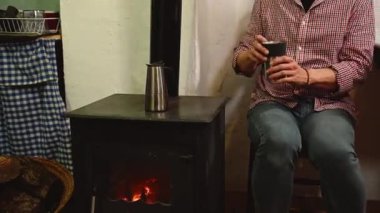Mavi kotlu ve kırmızı kareli gömlekli genç bir adam, paslanmaz çelik espresso makinesinden termos bardağına taze kahve dökerek, kırsal evdeki şöminenin yanında oturuyor.