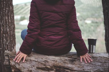 Ilık ceketli ve kot pantolonlu bir kadının, bahar ormanı erken saatlerinde oturduğu, termosunun yanındaki sıcak içecek bardağında kameraya döndüğü, güzel dağların manzarasına hayran kaldığı görüntüler.