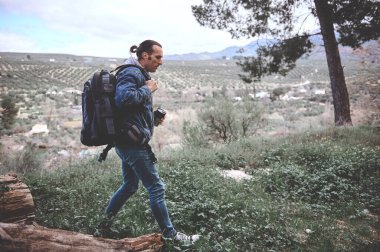Sırt çantası ve seyahat kupasıyla dağ manzaralı ormanda yürüyüş yapan genç bir yürüyüşçü. İlkbahar doğasını keşfeden, günlük kot giymiş beyaz bir gencin tam boy otantik portresi.
