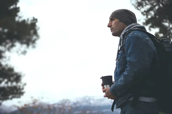 Side portrait of a male hiker adventurer with backpack, holding a thermos mug and dreamily looking away while hiking in the mountains of Sierra de Cazorla. Active people, trekking and travel concept