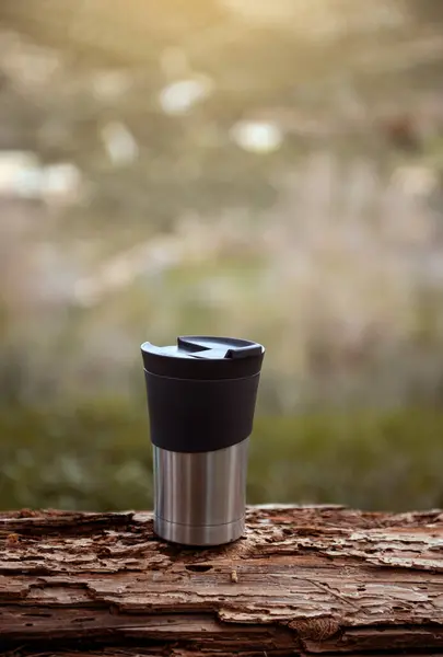 Vertical shot of a travel mug on a log over blur nature background with copy advertising space. A stainless steel cup with hot drink, coffee or tea for a recreation while trekking, hiking, travelling