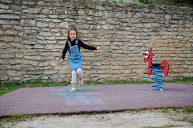 6-7 yaşlarında sportif bir kız çocuğu sırayla seksek oynuyor, okul bahçesinde işaretli meydanın üzerinden atlıyor. Klasik sokak çocuk oyunları işte. Çocukluk ve sağlıklı yaşam tarzı kavramı