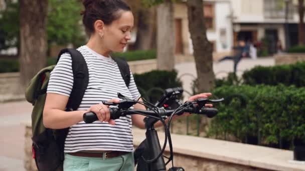 市内の電動自転車に乗っている間 カメラで笑顔の女性サイクリストの肖像画 バックパック付きのストライプTシャツの可愛い女性 携帯電話をチェック ダウンタウンの広場で彼女の自転車の近くに立って — ストック動画