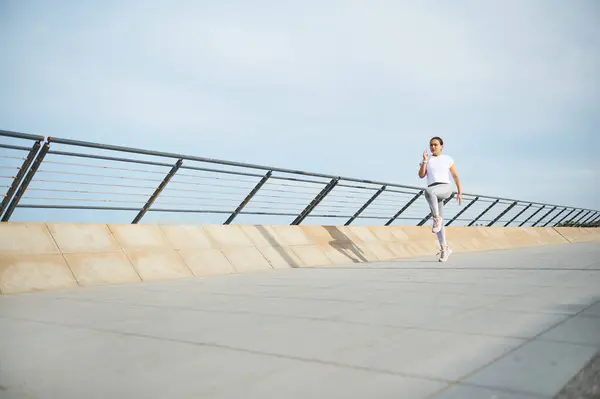 Full length portrait of a young athletic woman running in the city street. Happy female runner burning calories, performing morning jog on the promenade. People. Sport. Active healthy lifestyle.