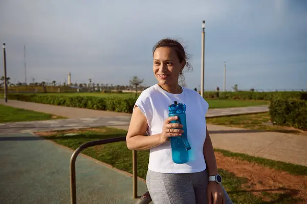Confident woman in white t-shirt, determined young athlete, sportswoman holding bottle of water, smiling looking side, relaxing after heavy bodyweight training or outdoor workout on urban sportsground