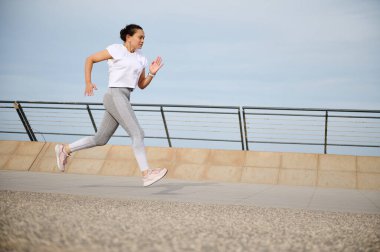 Şehir köprüsünde koşan bir kadın. Aktif sporcu bayan sporcu sabah antrenmanından, koşudan hoşlanır. Sağlıklı yaşam tarzı kavramı. Spor kıyafetli, açık havada egzersiz yapan, beyaz tişörtlü bir sporcu.