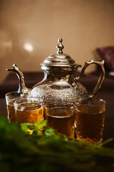 Close-up drinking glasses with fresh Moroccan tea with mint and silver traditional tea pot on wooden table. A tea break with traditional the a la menthe as symbol of Moroccan hospitality