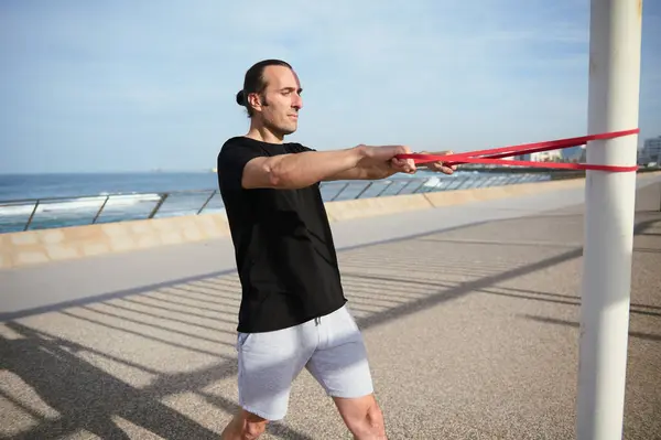Determined young man with athletic physique performing bodyweight training, using a resistance fitness band. Sportsman doing exercises on shoulders on the promenade outdoors. Copy advertising space.