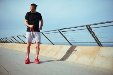 Uzun boylu sporcu, siyah tişörtlü atletik adam, plajda bir direnç fitness bandıyla pazılar üzerinde egzersiz yapıyor. Kararlı erkek sporcu şehirde vücut ağırlığı antrenmanı yapıyor.