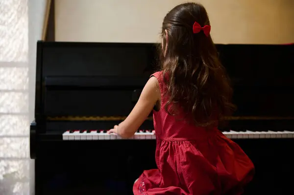 Rear view of a cute child girl, a pianist student, future musician pianist touching the white keys with his fingers to create the rhythm of the melody, enjoying playing grand piano during music lesson