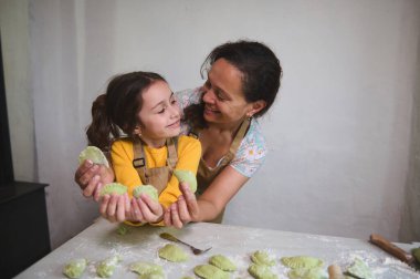 Gülümseyen anne ve onun mutlu kızı birbirlerine bakıyor, el yapımı heykel ya da Ukraynalı varenky, un kaplı mutfak masasında yan yana duruyor, beyaz duvar arka planına karşı