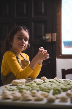 Kafkasyalı güzel bir kız çocuğu, bej önlüklü küçük bir şef, ev yapımı bir hamur köftesi tutuyor, kameraya bakıp gülümsüyor, un kaplı masada mantı, pelmeni, vareniki, hinkali ile duruyor.