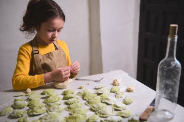 Beyazlar içinde bej rengi şef önlüğü takmış, hamur köftelerini kalıba döküyor, un kaplı mutfak masasında duruyor. Küçük kız, annesine evde yemek pişirmesinde yardım ediyor.