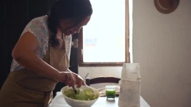 Mutfakta tahta kaşıkla hamur yoğurmayı seven hoş bir ev hanımı. Bej mutfak önlüğüyle kadın aşçı pastası kasede malzemeleri karıştırıyor, hamur köftesini patates püresiyle hazırlıyor.