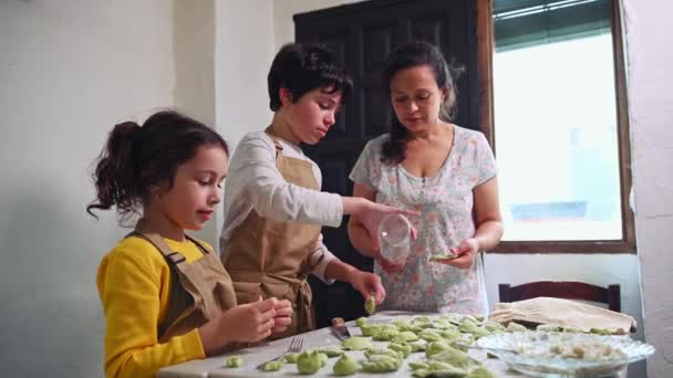 ベージュのシェフエプロンの素敵な子供たち フラワーキッチンテーブルに立って 彼らの母親が詰め物や彫刻を助けます 娘と息子は伝統的なレシピに従って一緒に料理する — ストック動画