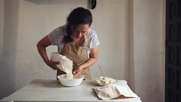 ベージュエプロンで快適な主婦 キッチンテーブルに立って 白いボウルにいくつかの全体の穀物小麦粉を注ぐ 素朴なキッチンインテリアの大理石の表面のダンプリングのための生地を準備 — ストック動画