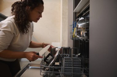 Otantik ev kadınının mutfak malzemelerini temizleyip, aşırı yüklenmiş bulaşık makinesinden sterilize edişinin yan görüntüsü. Ev işleri için bulaşık makinesi kullanan güzel bir kadın. Mutfak aletleri.