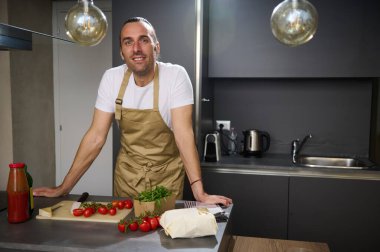 Kafkasyalı yetişkin bir adamın otantik portresi, bej önlüklü bir şef, mutfak tezgahında taze malzemelerle duruyor, kameraya güvenle bakıyor, akşam yemeği için sağlıklı salata pişiriyor.