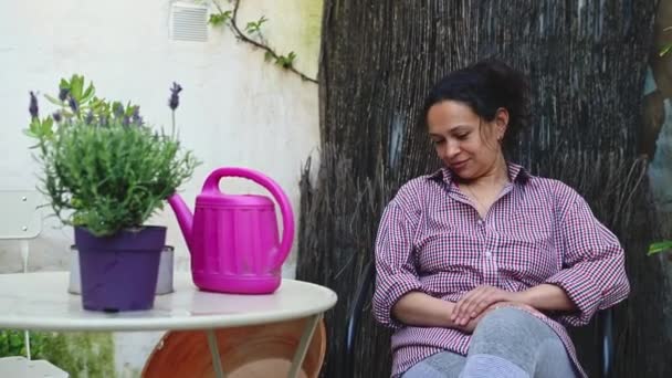 ハッピーな女性が家の庭の椅子に座り 仕事の後にリラックスしています テーブルの上の水やり場の近くで咲くラベンダーの焦点の変更 田舎の環境で追い出される若い女性 — ストック動画