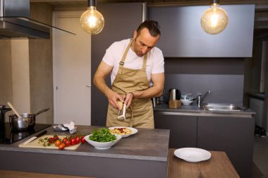 Yakışıklı beyaz bir adam, bej önlüklü bir erkek şef, domates soslu bir spagetti içinde parmesan peyniri rendeliyor, ev mutfağında geleneksel İtalyan tarifine göre akşam yemeği pişiriyor..