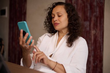 Evde cep telefonu kullanan güzel bir kadın, haberlere kayıyor, mobil uygulamaları kontrol ediyor, haberleri okuyor, online alışveriş yapıyor, otel planlama tatil seyahatleri düzenliyor, rahat bir evde mutfak masasında oturuyor.