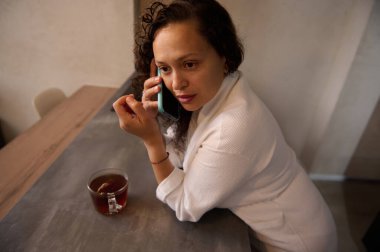 Genç çok ırklı bir kadın yana bakıyor, kahvaltısını evde yaparken akıllı cep telefonuyla konuşuyor, evde mutfak tezgahında beyaz bornozla dikiliyor. Yakın plan yan portre