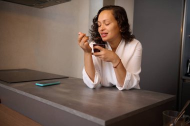 Gülümseyen güzel kadın kahvaltıda çikolatalı yoğurt yiyor, mutfak masasında duruyor.