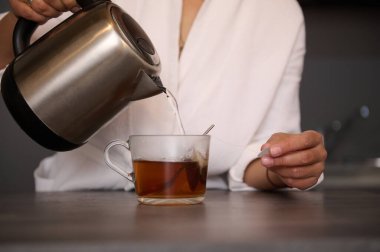Kahvaltı için çay yapan bir kadının yakın çekimi. Seçici olarak, elektrikli paslanmaz çelikten çay poşeti ile cam fincana kaynar su döken kadın ellerine odaklan. Yiyecek ve içecek tüketimi