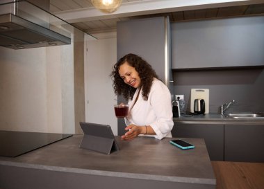 Kahvaltıda sıcak çay içen ve dijital tabletle video bağlantısıyla konuşan, mutfak tezgahında beyaz bornoz giymiş çok ahlaklı mutlu bir kadın.