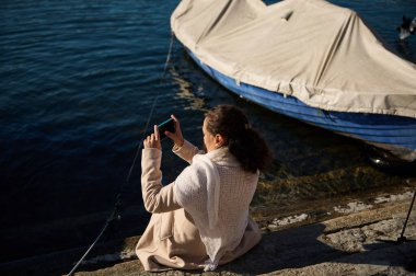 Bej ceketli, Como Gölü kıyısında teknelerin yanında oturan ve akıllı cep telefonuyla manzarayı fotoğraflayan genç bir kadının arka görüntüsü. Güzel bayan turist göl kenarında dinleniyor.