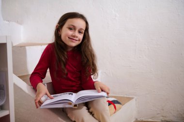 Zeki ilkokul öğrencisi kız elinde kitapla evde oturmuş kameraya gülümsüyor. Sevimli çocuk kitabı okuyor. Dünya Kitap Günü konsepti. Akademik yılın yeni döneminde okula dönüyorum.