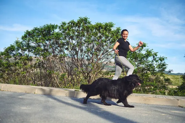 Açık havada köpeğini tasmayla gezdiren ve koşan melez bir genç kadının tam boy portresi. - İnsanlar. Evcil hayvanları oynuyorum. Spor. Aktif ve sağlıklı yaşam tarzı kavramı