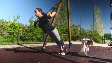 40 'lı yaşlarda orta yaşlı atletik bir kadının tam boy yan portresi, askı kayışlarıyla kol egzersizleri yapıyor, açık hava kentsel spor sahasında çalışıyor. Halk ve spor