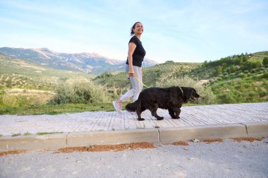 Aktif giyinmiş bir kadının dağlarda köpeğini gezdirirken çekilmiş uzun bir resmi. Evcil hayvanlar, hayvanlar ve insanlar konsepti