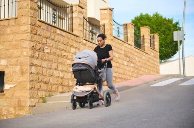 Siyah tişörtlü, gri spor taytlı, dışarıda egzersiz yaparken bebek arabasını iten gülümseyen genç bir kadın. - İnsanlar. Annelik ve bebeklik. Bebekli fitness. Aktif ve sağlıklı yaşam tarzı kavramı