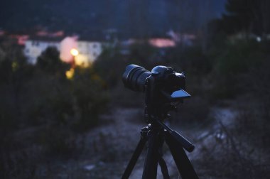 Bir gezgin fotoğrafçı tarafından tripoda yerleştirilmiş siyah dokunmatik ekranlı dijital bir kamera, gün doğumunda dağlardaki güzel bir şehrin görüntüsünü yakalıyor. Dünya Fotoğraf Günü konsepti