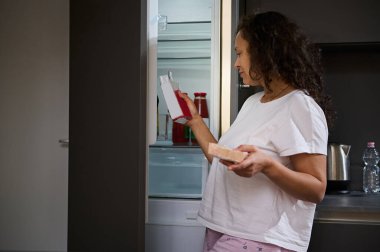 Açık bir buzdolabının yanında duran güzel bir kadının özgün portresi ellerinde yiyecek paketlerine bakıyor, mutfağında kahvaltı hazırlıyor. - İnsanlar. Yiyecek ve içecek tüketimi.