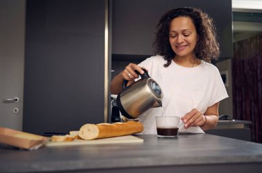 Gülümseyen Latin Amerikalı kadın evde, kahve yapıyor. Güzel esmer ve beyaz pijamalı, mutfak masasında duruyor, kahvaltı hazırlıyor, çaydanlıktan suyu bardağa dolduruyor ve sandviç yapıyor.