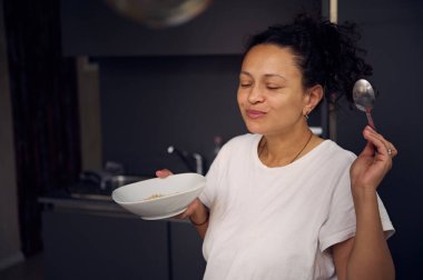 Güzel kıvırcık saçlı esmer, pijamalı genç bir kadın, ev mutfağında duruyor, sabah kahvaltısında sağlıklı yulaf gevreğinin tadını çıkarıyor. - İnsanlar. Yaşam tarzı. Gıda tüketimi.